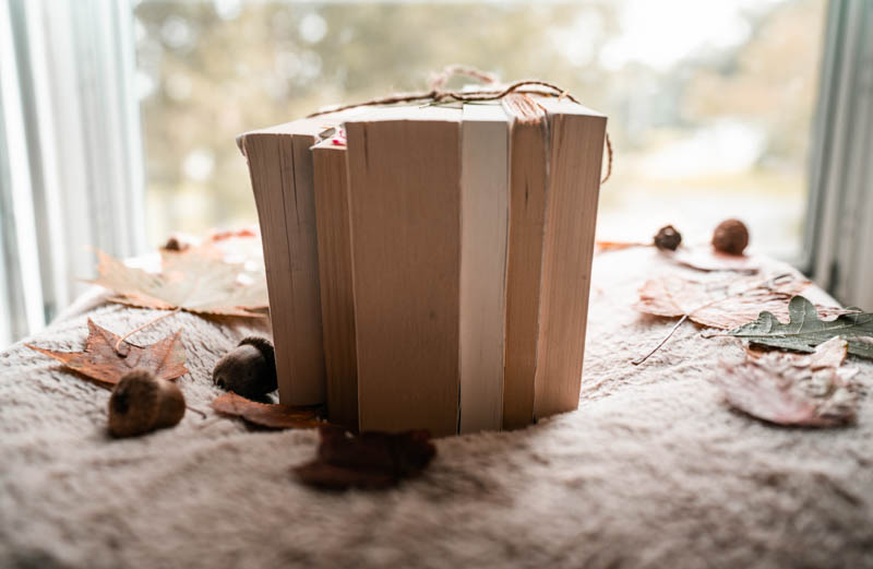 A stack of 5 books wrapped in twine sitting on the window surrounded by autumn leaves and acorns.