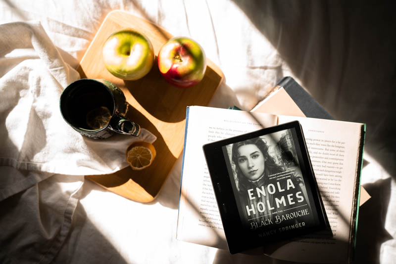 My Kindle Oasis opened to the cover of Enola Holmnes and the Black Bourrache next to some of my hand picked authumn apples and hand crafted (from a friend) coffee mug.